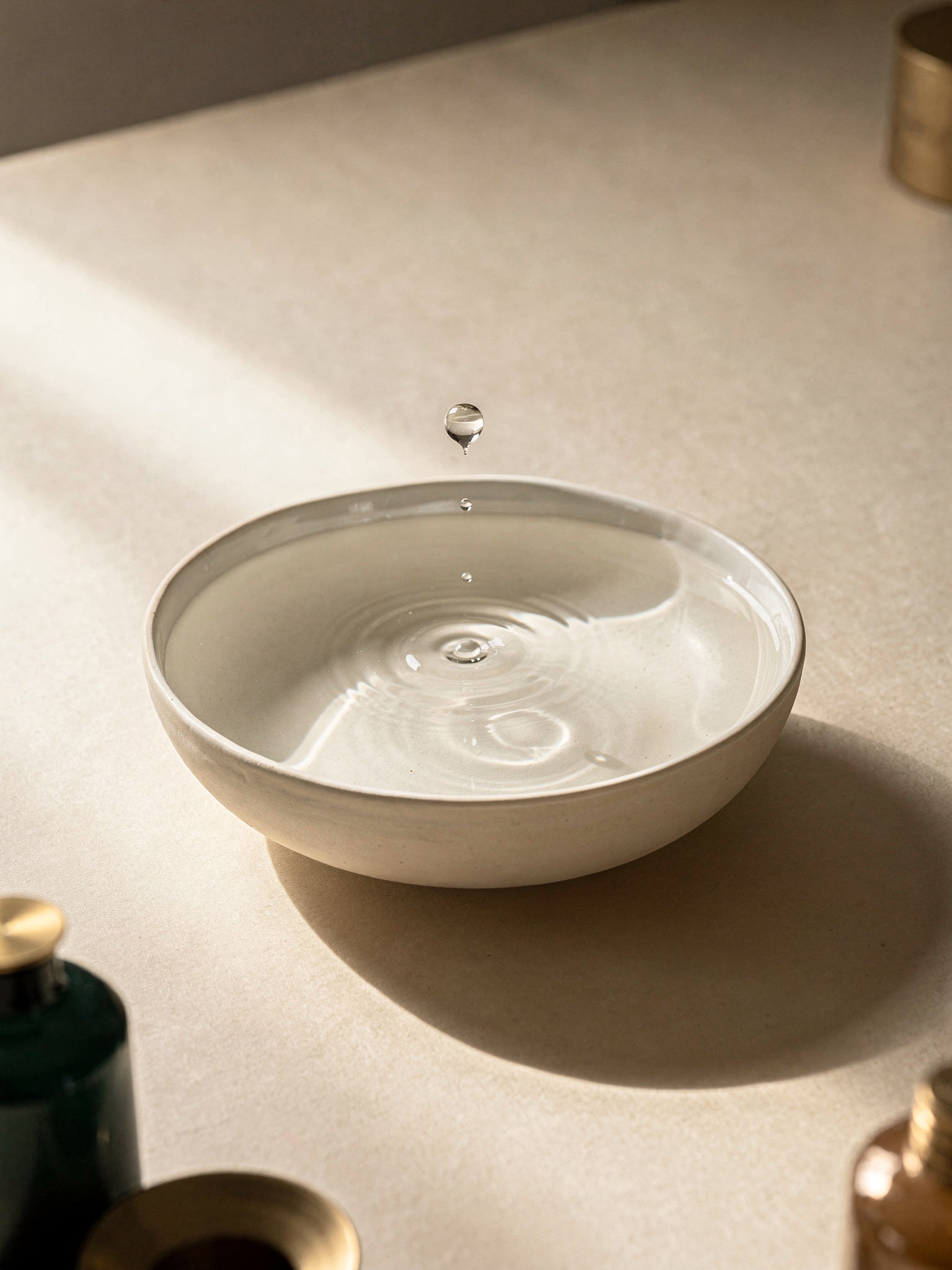 A pale ceramic bowl of perfectly still water with a single drop suspended above creating one concentric ripple.